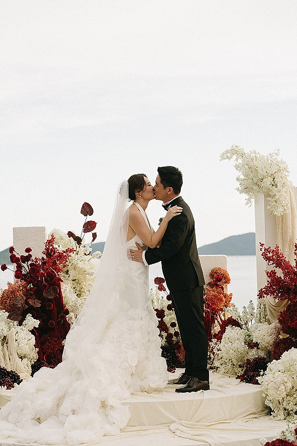 Wedding kiss at the altar as bride in cathedral veil and groom in black tuxedo embrace beneath a floral arch by the water and hills