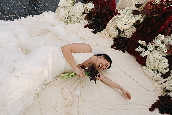 Bridal portrait of a bride lying on patterned stone pavers, laughing in a strapless tulle gown with a calla lily bouquet and burgundy florals