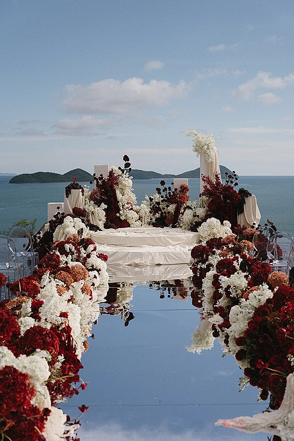Ceremony aisle decor with mirrored wedding aisle lined in red and white florals and draped fabric, set by the ocean and islands under blue sky