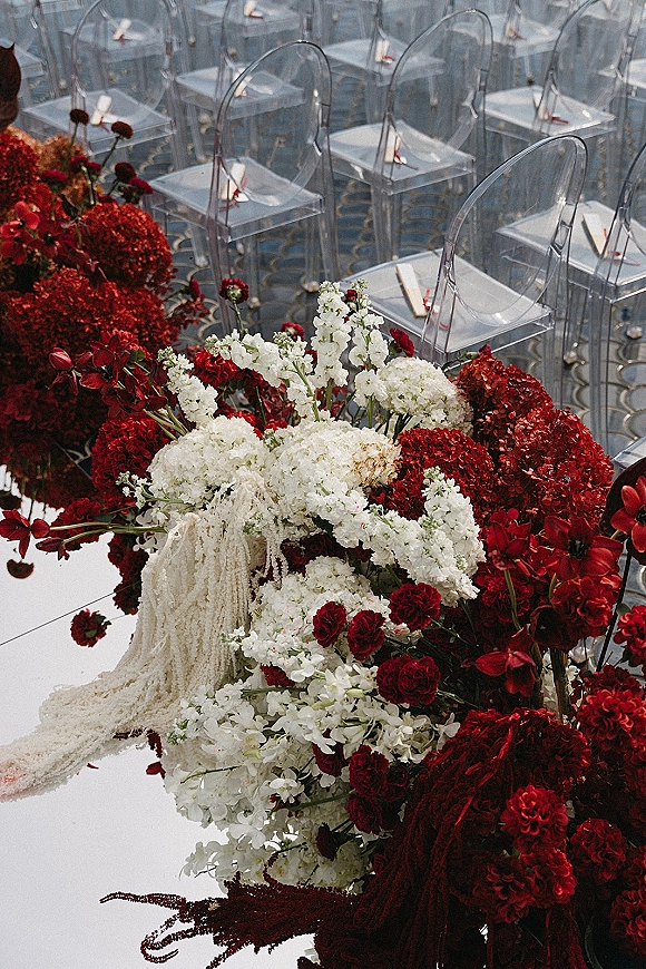 Ceremony aisle flowers in a wedding aisle floral arrangement of red and white hydrangeas, roses and tulips beside acrylic chairs on patterned carpet indoors