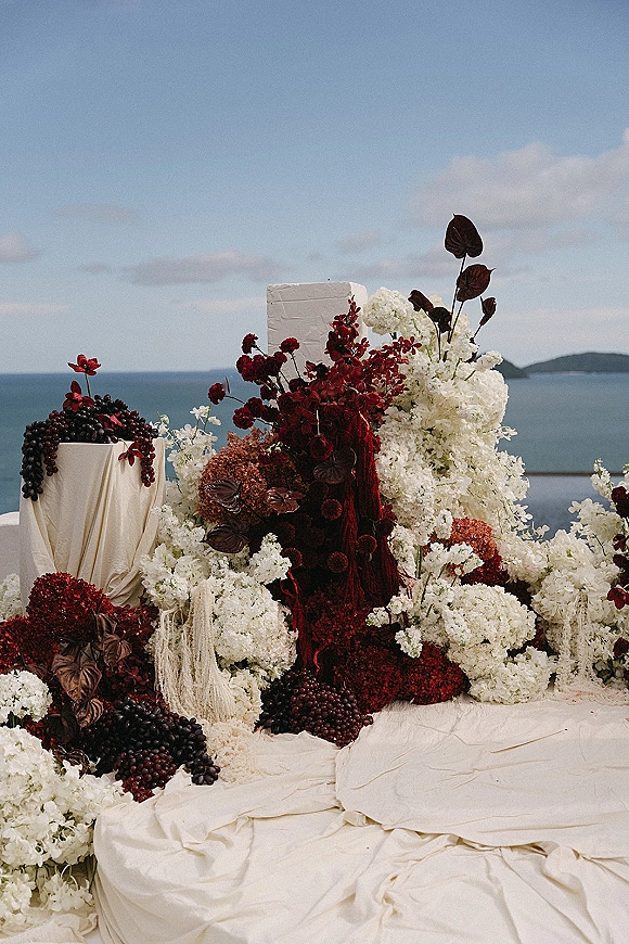 Wedding ceremony backdrop with ceremony floral installation of burgundy and white flowers, draped fabric, and plinths overlooking the ocean view