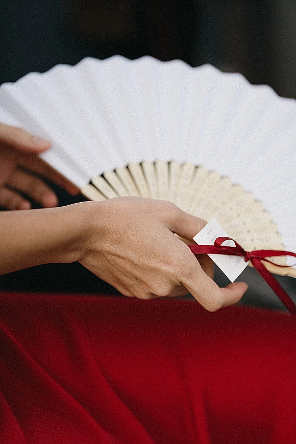 Wedding hand fans with a ceremony program fan design, tied with a red ribbon and name tag against a dark backdrop with red fabric