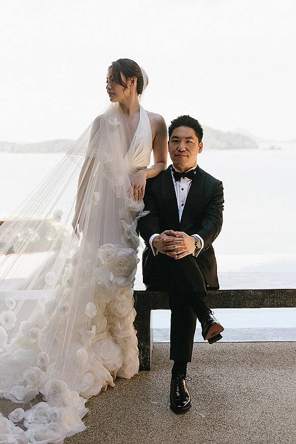 Couple portrait of bride leaning on seated groom, veil blowing, on an ocean terrace with stone railing and distant hills behind