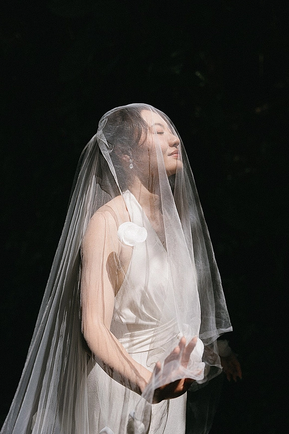 Bridal portrait of a bride wearing veil over her face, in a white gown with pearl drop earrings against a dark background