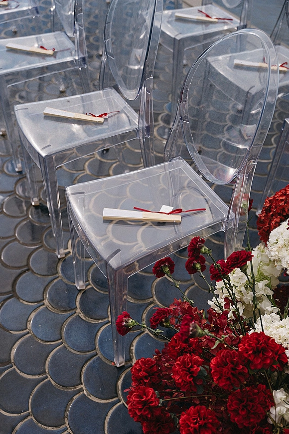 Ceremony aisle setup with clear acrylic wedding chairs, programs tied with red ribbon, and red-white florals on a patterned tile floor
