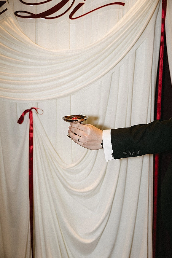 Wedding cocktail in a martini glass with cherry garnish held by groom in suit, wedding band and cufflinks against white draped wall