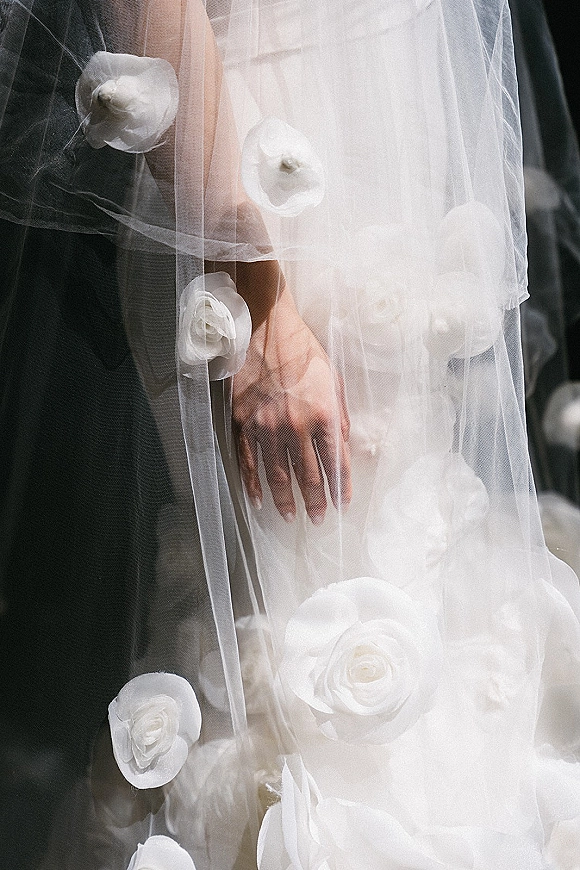 Wedding veil detail with 3D floral veil appliques on textured tulle, bride’s hand smoothing the dress beside dark suit fabric indoors