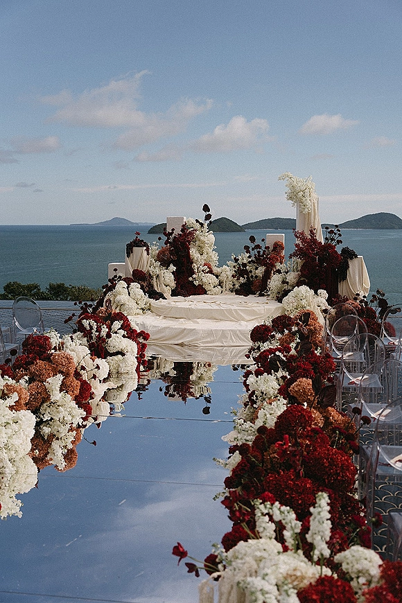 Ceremony aisle design with a mirrored wedding aisle runner, floral-lined edges, draped fabric, and clear acrylic chairs on an ocean-view terrace