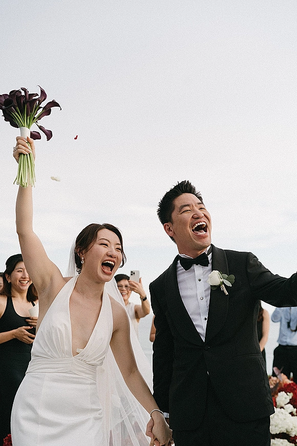 Wedding recessional as newlyweds cheering, holding hands while bride lifts a calla lily bouquet, veil flowing past guests outdoors under open sky