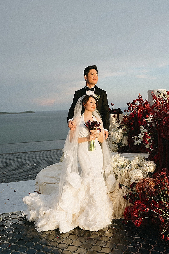 Couple portrait of bride and groom, her cathedral veil flowing as she holds a bouquet on a stone terrace overlooking the ocean horizon