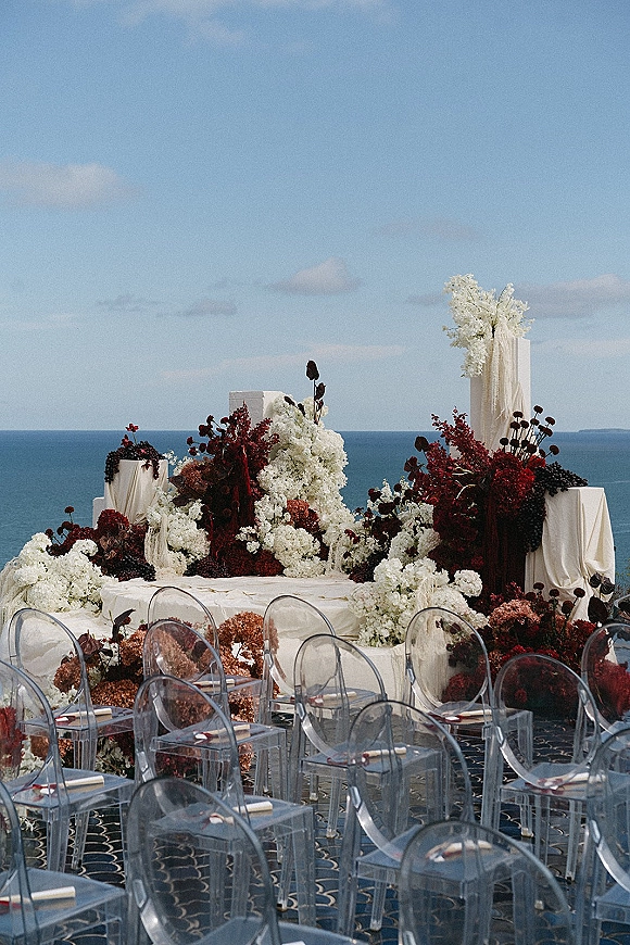 Ceremony setup with oceanfront wedding ceremony florals, draped fabric, clear acrylic chairs, and aisle runner on a terrace with ocean view