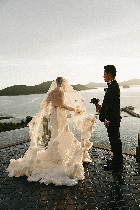 Couple portrait at an infinity pool, bride and groom silhouette facing each other, veil and dress train flowing over the terrace at sunset