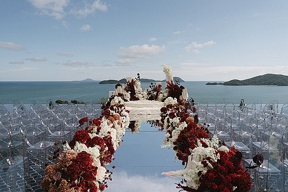 Ceremony aisle design with a mirror wedding aisle runner, red and white floral pillars and ghost chairs on a terrace with ocean view