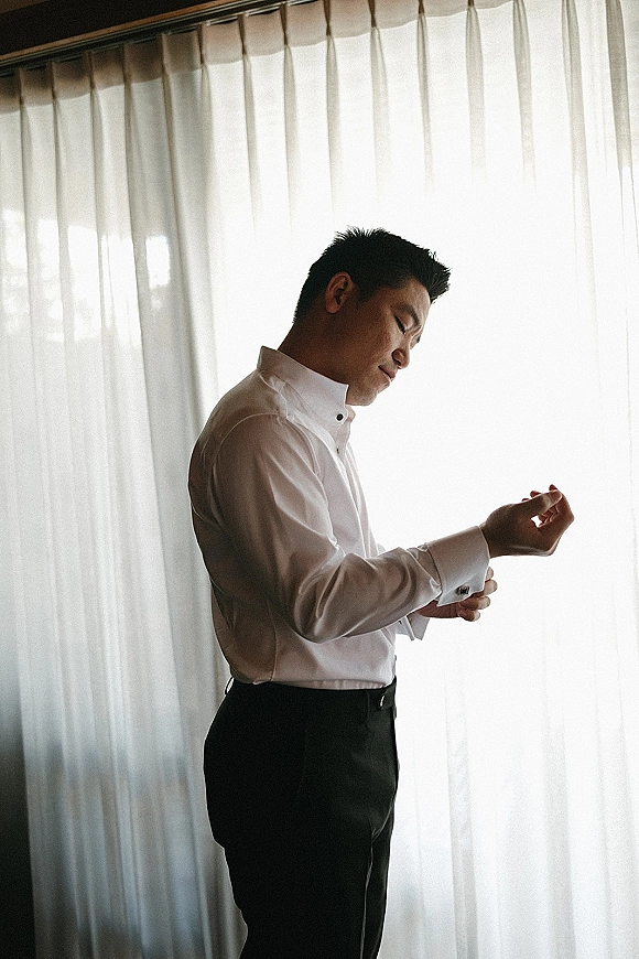 Groom getting ready, groom putting on cufflinks on a white dress shirt, standing by sheer curtains in soft window light