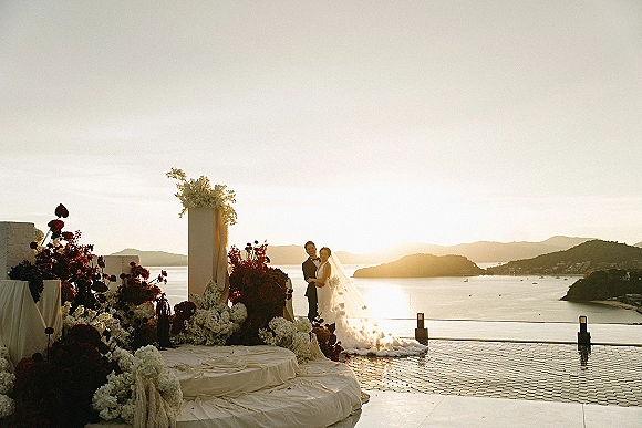 Couple portrait at a sunset wedding portrait, bride in veil and groom in tuxedo embracing on a terrace with ocean and mountains behind
