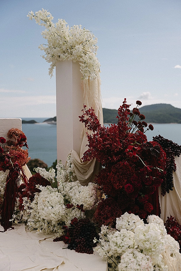 Ceremony altar decor with wedding altar flowers on white column pedestals, draped fabric, and burgundy blooms, set against ocean and mountains backdrop
