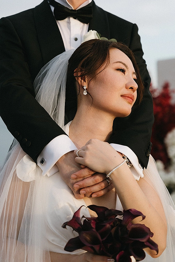 Couple portrait of groom hugging bride in strapless dress and veil, holding a burgundy bouquet against soft greenery and sky backdrop