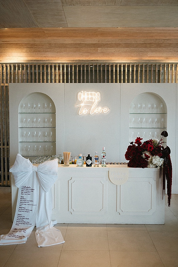 Wedding bar setup with neon sign above a white counter, glassware and liquor display, cocktail menu, and floral arrangement on arched shelves
