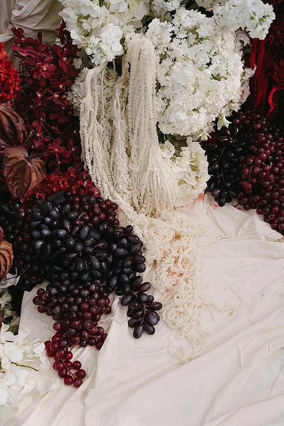 Wedding tablescape with fruit wedding centerpiece featuring white hydrangeas, burgundy blooms, black and red grapes on cream draped fabric and beaded fringe runner