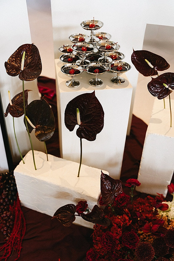 Champagne tower setup with a coupe glass tower of dark red cherry-garnished drinks on white pedestals with anthurium flowers indoors