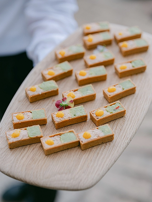 Wedding dessert bites and wedding petit fours on a wood serving board with edible flower garnish, held by a catering server on neutral floor
