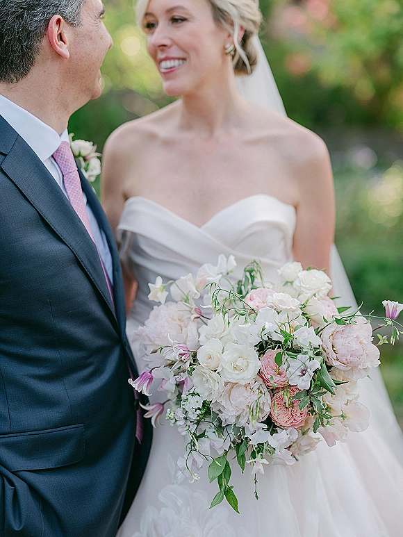 Couple portrait of bride in strapless dress and veil holding a blush-and-white bouquet beside groom in navy suit with pink tie, garden bokeh behind