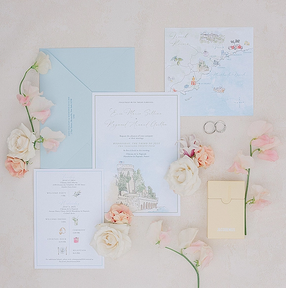 Invitation suite wedding stationery flat lay with pastel blue envelope, map card, rings and blush flowers on light fabric surface