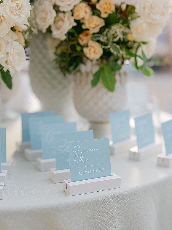 Wedding place cards and blue escort cards with calligraphy lettering in white holders on a linen table with floral vases and greenery accents