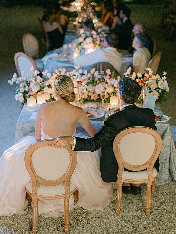 Reception moment at a candlelit sweetheart table decor setup with floral centerpiece, champagne flutes, and dimly lit guests behind