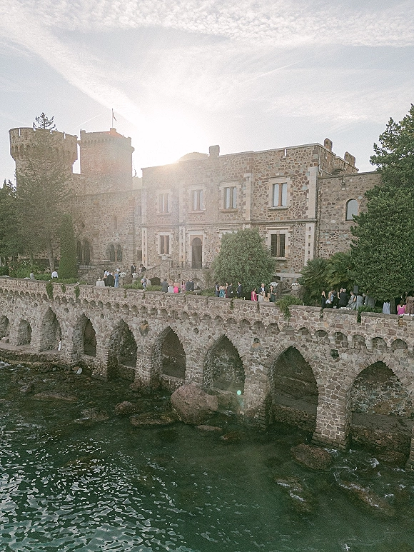 Wedding venue exterior with a castle wedding venue stone facade and arched bridge walkway overlooking ocean water and rocky shoreline in sunlight