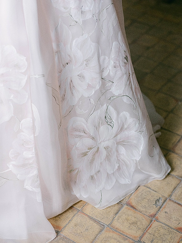 Wedding dress detail with 3d floral wedding dress appliqué and sheer embroidery overlay, the hem resting on a tile floor