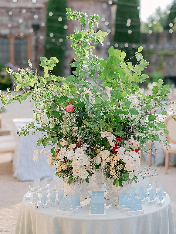 Escort card display with wedding escort cards in holders on a round table, topped by a tall blush floral arrangement under patio string lights