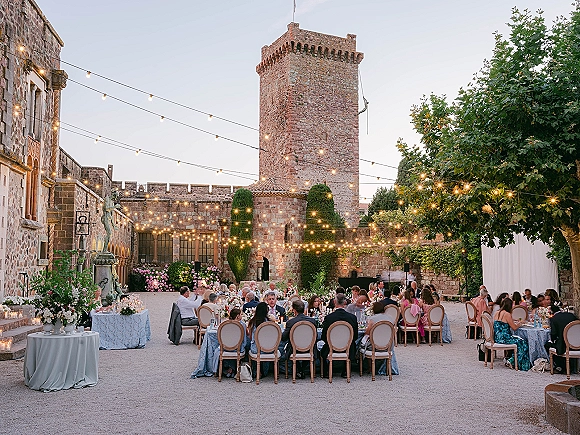 Outdoor reception decor with string lights over long banquet and round tables, blue linens, candles, florals, and a fountain in a castle courtyard