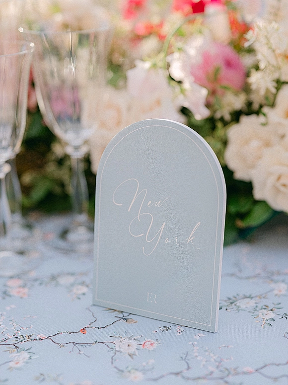 Wedding place card with arched place card design and calligraphy lettering on a patterned tablecloth beside champagne flutes and flowers