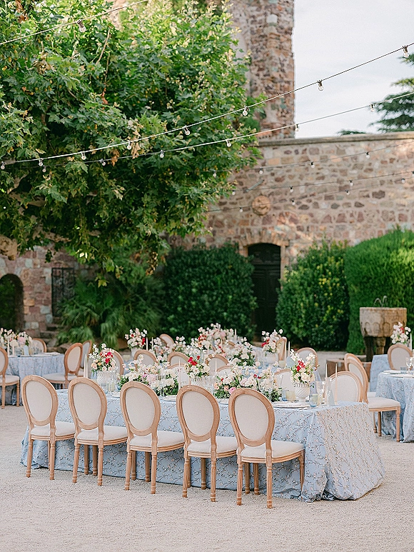 Reception tablescape with blue patterned tablecloths and floral centerpieces, round tables with candles and glassware under string lights in a stone courtyard