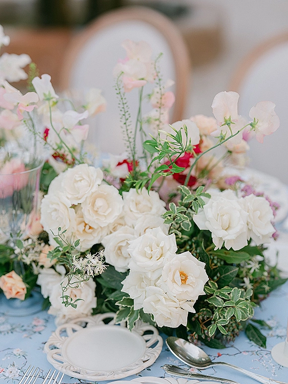 Reception tablescape with wedding floral centerpiece of white roses, pastel sweet peas and greenery on patterned tablecloth, neutral seating behind