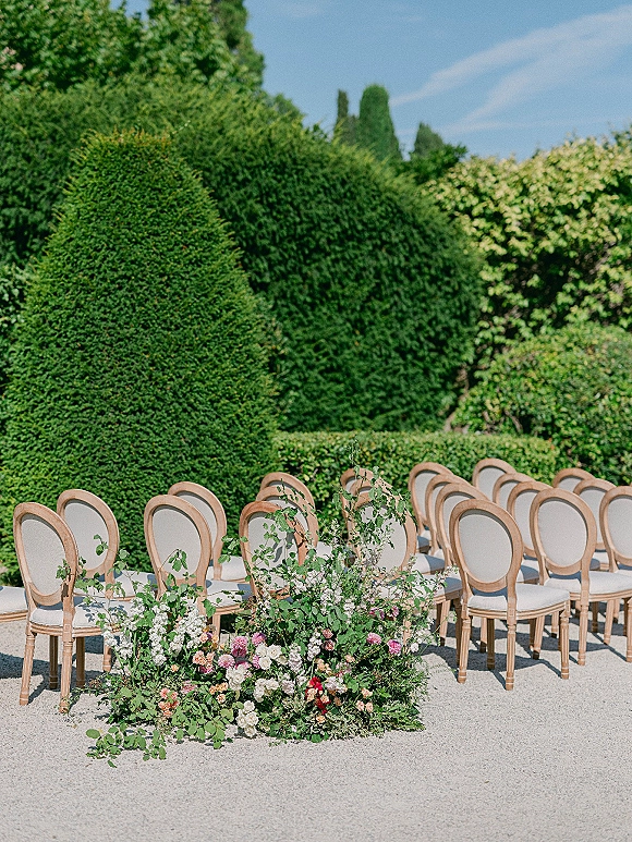 Ceremony aisle decor with outdoor ceremony seating, wood and upholstered chairs flanked by low floral greenery on a gravel path by hedges