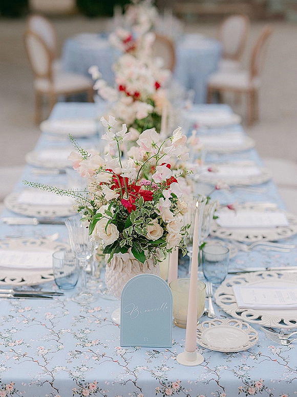 Reception tablescape with blue floral tablescape linens, floral runner centerpiece, pink taper candles, menu cards, and blue glass goblets on a long table