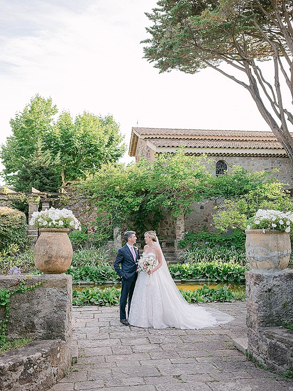 Couple portrait of bride and groom portrait with bride holding a white bouquet, veil and navy suit, on a stone walkway by urn planters