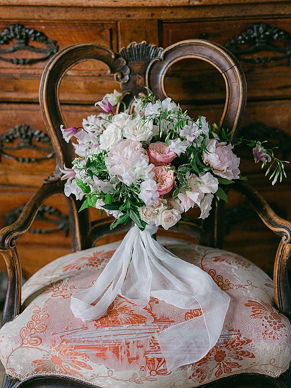 Wedding bouquet of blush and white garden roses with greenery and a long white ribbon streamer resting on an upholstered vintage chair