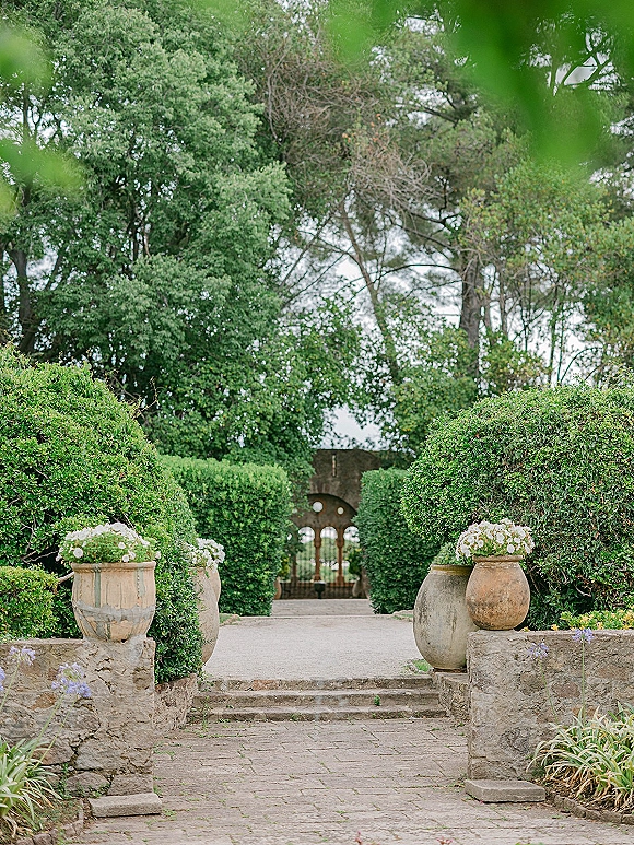 Garden walkway at a wedding venue entrance with hedges and stone planters of white flowers leading up steps to a wrought iron gate