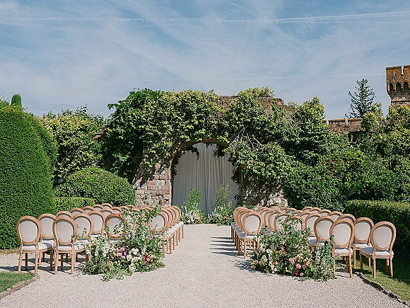 Outdoor ceremony setup with garden wedding ceremony aisle florals lining a gravel path to a stone archway with draped fabric and vines under blue sky