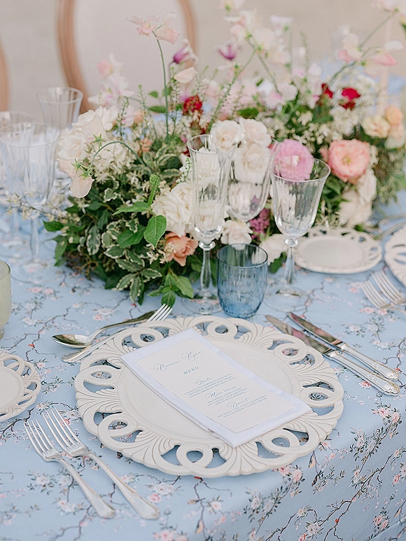 Reception tablescape with a wedding table centerpiece of roses and greenery, blue glass tumbler, champagne flutes, menu card, and cutwork charger plate