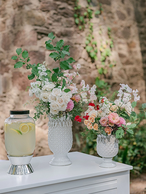 Wedding drink station with a glass beverage dispenser of lemon and lime water on a white cabinet, set against an ivy-covered stone wall