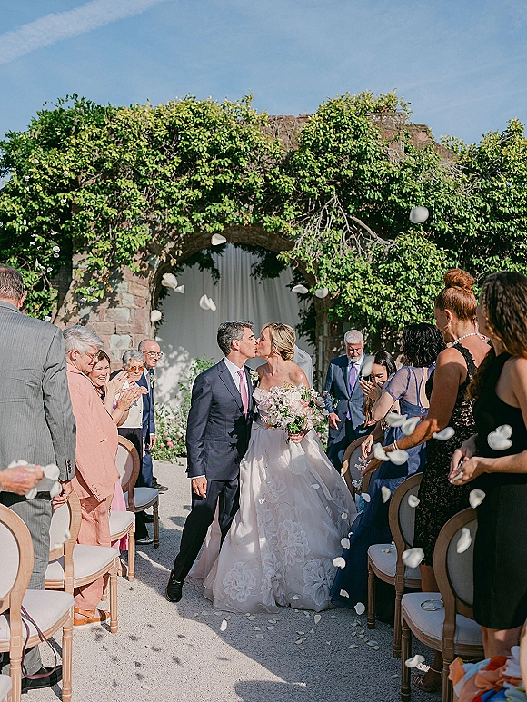 Wedding kiss as newlyweds walk down the aisle, bride in veil holding bouquet while guests toss petals under a stone archway