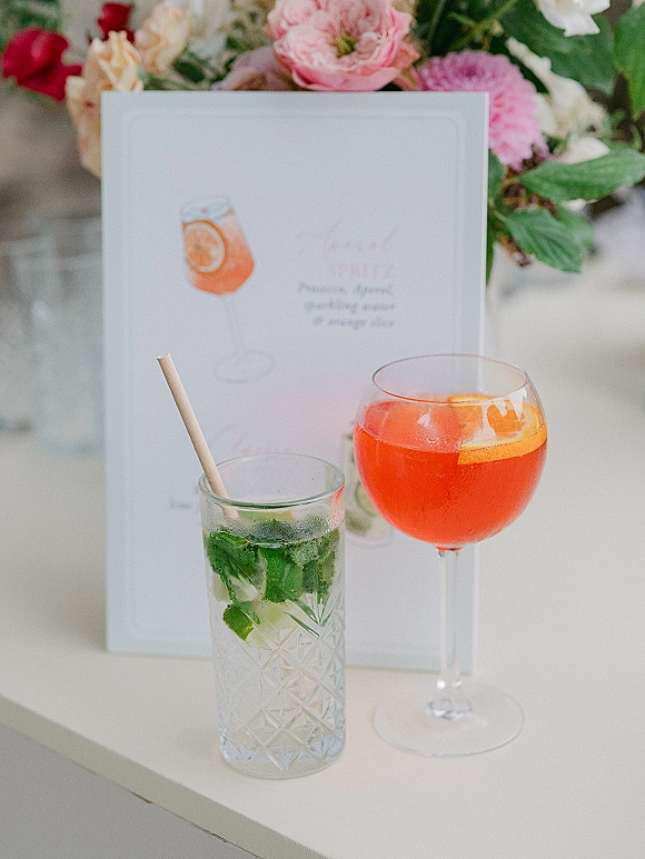 Wedding signature drinks beside a signature cocktail menu sign, goblet and textured highball glasses with citrus, mint, ice on white table