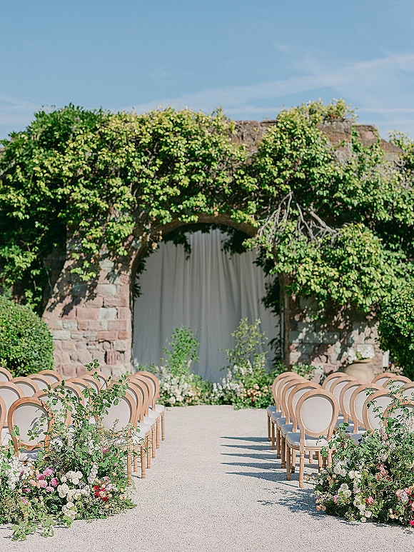 Ceremony setup with wood chairs and white cushions beside a floral lined aisle leading to a draped backdrop under a stone wall arch