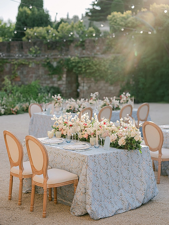 Reception tablescape with light blue floral tablecloth, blush and white centerpieces, taper candles, and place cards in an ivy courtyard under string lights