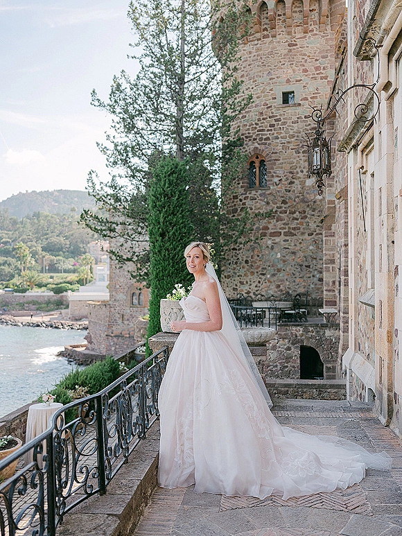 Bridal portrait of a bride in a strapless gown and cathedral veil holding a bouquet on a stone castle terrace overlooking the coast