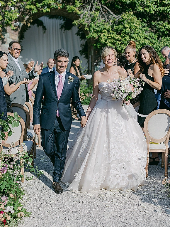 Wedding recessional as bride and groom walk the aisle hand in hand, she holds a blush bouquet, guests cheer in a garden courtyard with stone archway